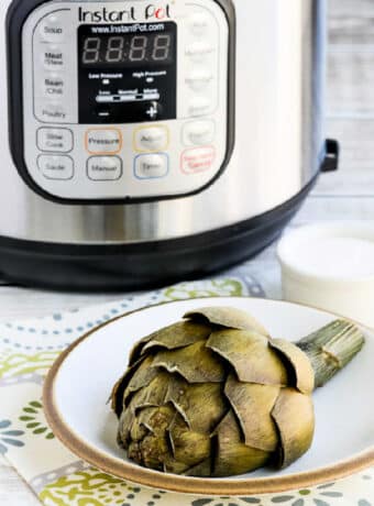 Square image of artichoke on plate and Instant Pot in back for Instant Pot Artichokes.
