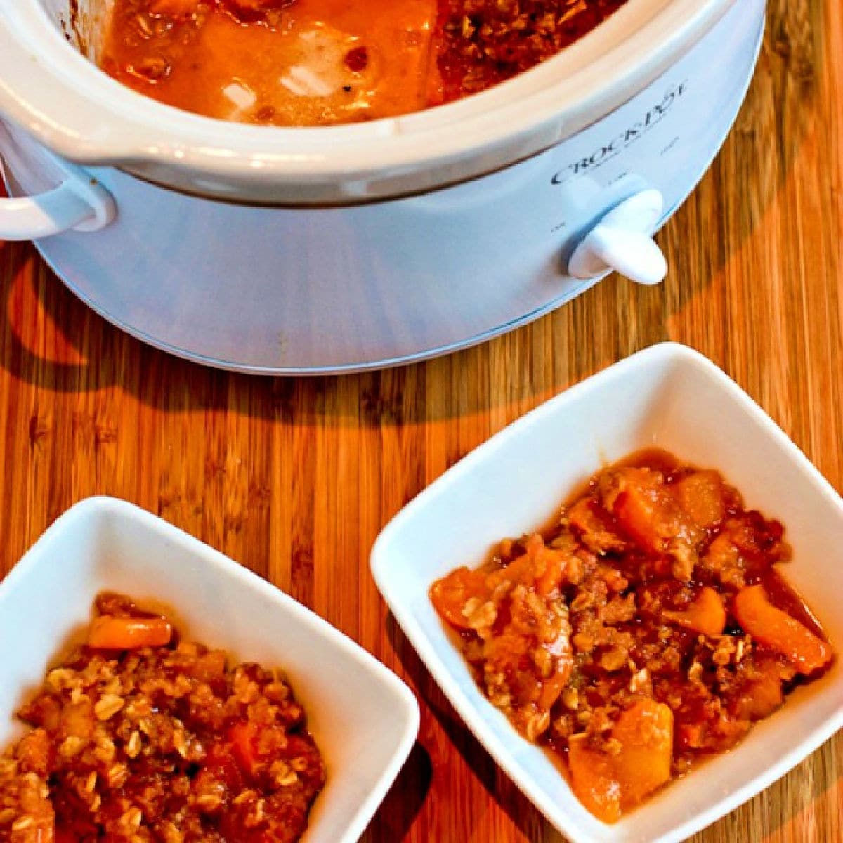 Square image of Slow Cooker Peach Crisp shown in two serving bowls with slow cooker in back.