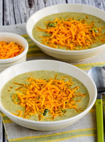 Cheesy Broccoli and Cauliflower Soup shown in two serving bowls with extra cheese on top and on the side.