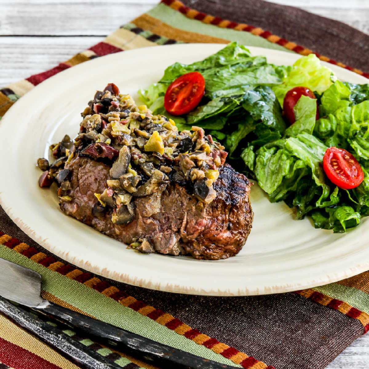 Square image for Pan-Grilled Steak with Olive Sauce shown on plate with lettuce and tomato salad.