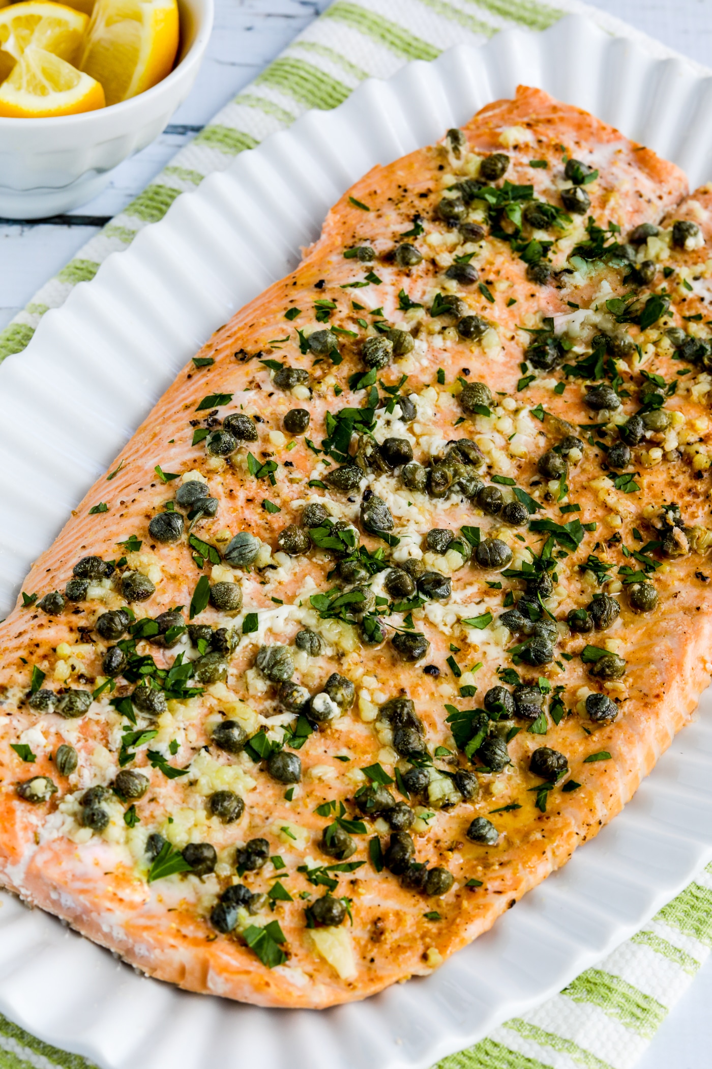 large salmon fillet on serving plate with butter, garlic, capers, and lemon