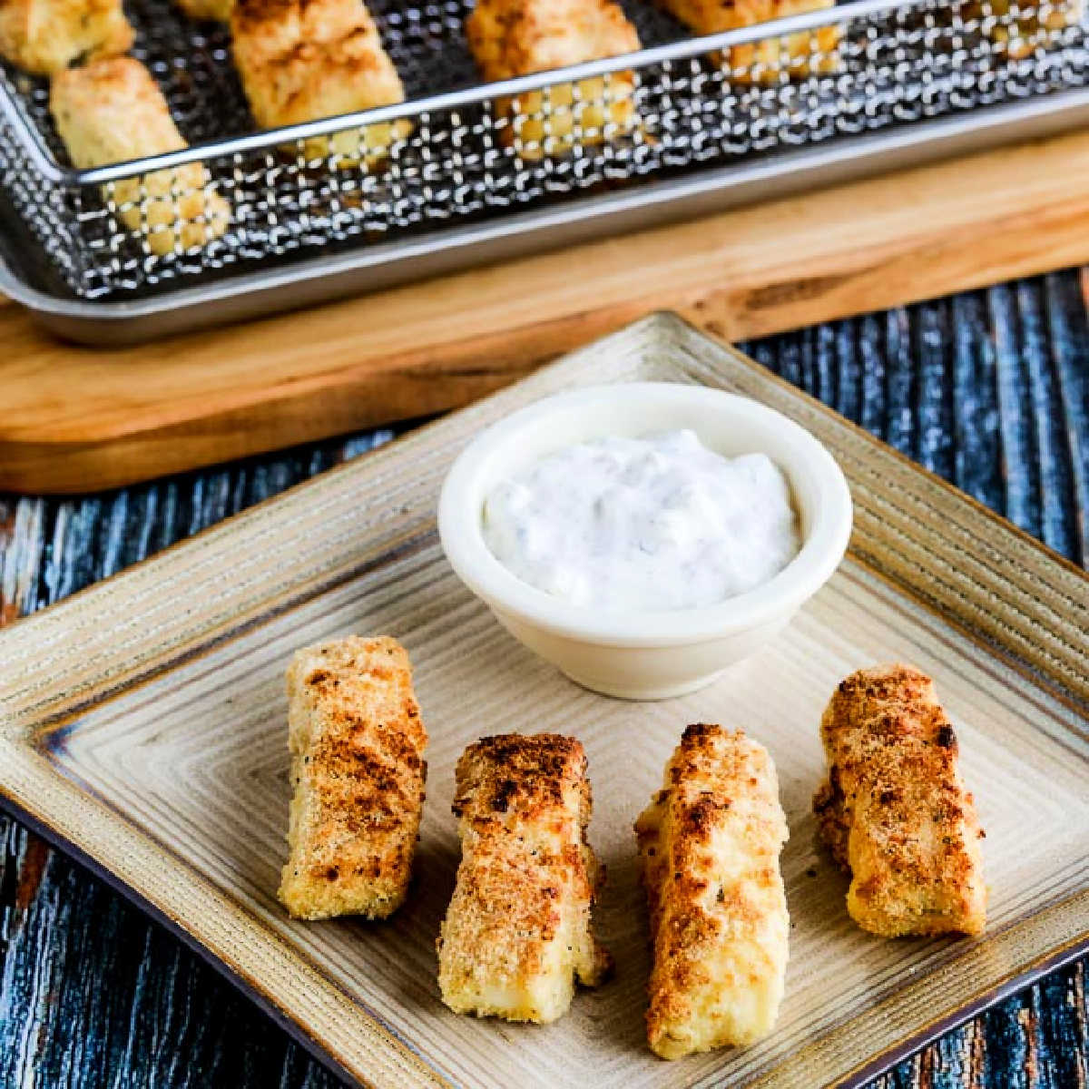 square image of Air Fryer Fish Sticks on serving plate with tartar sauce, air fryer in background.