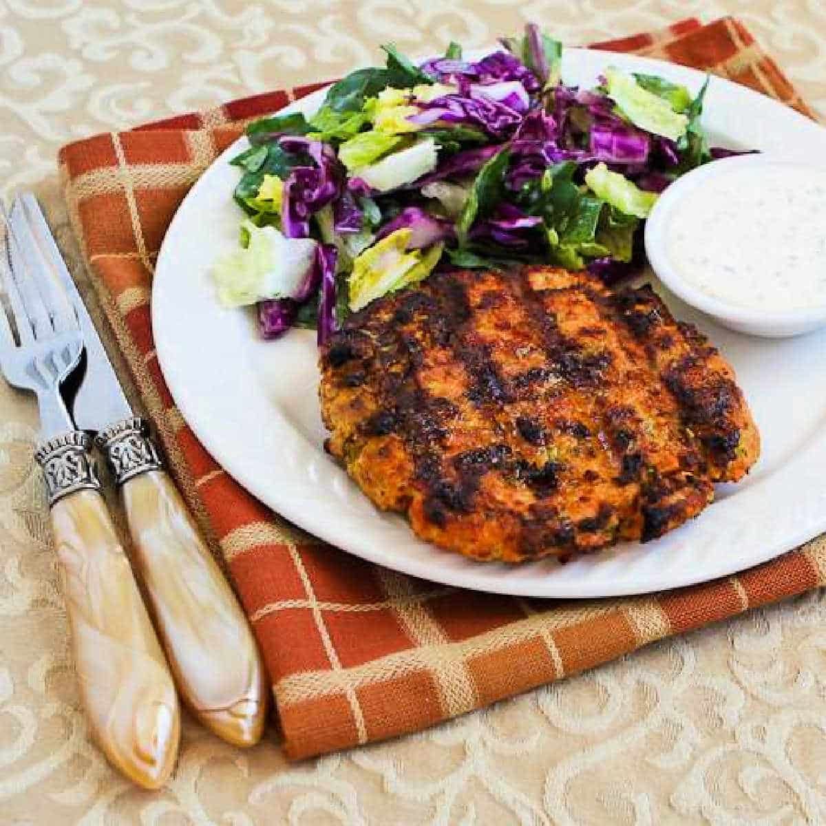 Square image of Grilled Salmon Burgers with Caper Mayo shown on serving plate with salad, on napkin.