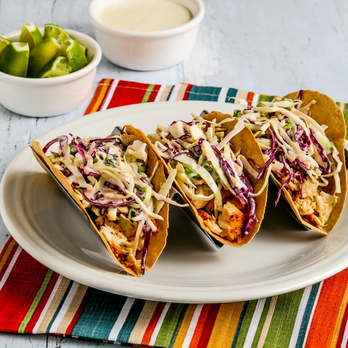 Square image of air fryer fish tacos, tacos with lime and additional sauce on a taco rack.