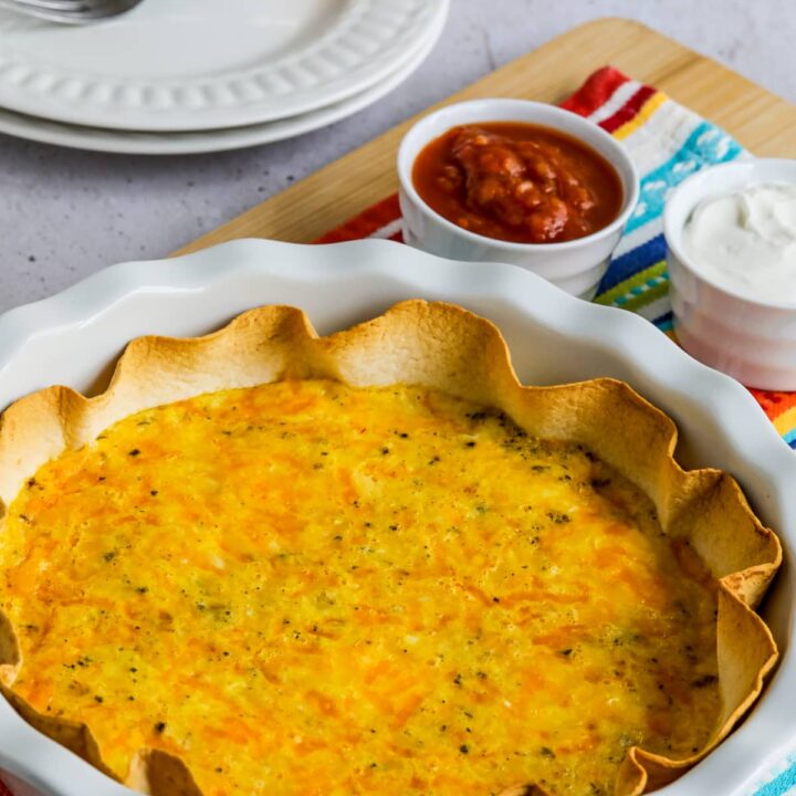 Egg and Cheese Tortilla Bake shown in white pie dish with sour cream and salsa on the side and plates/forks in back.
