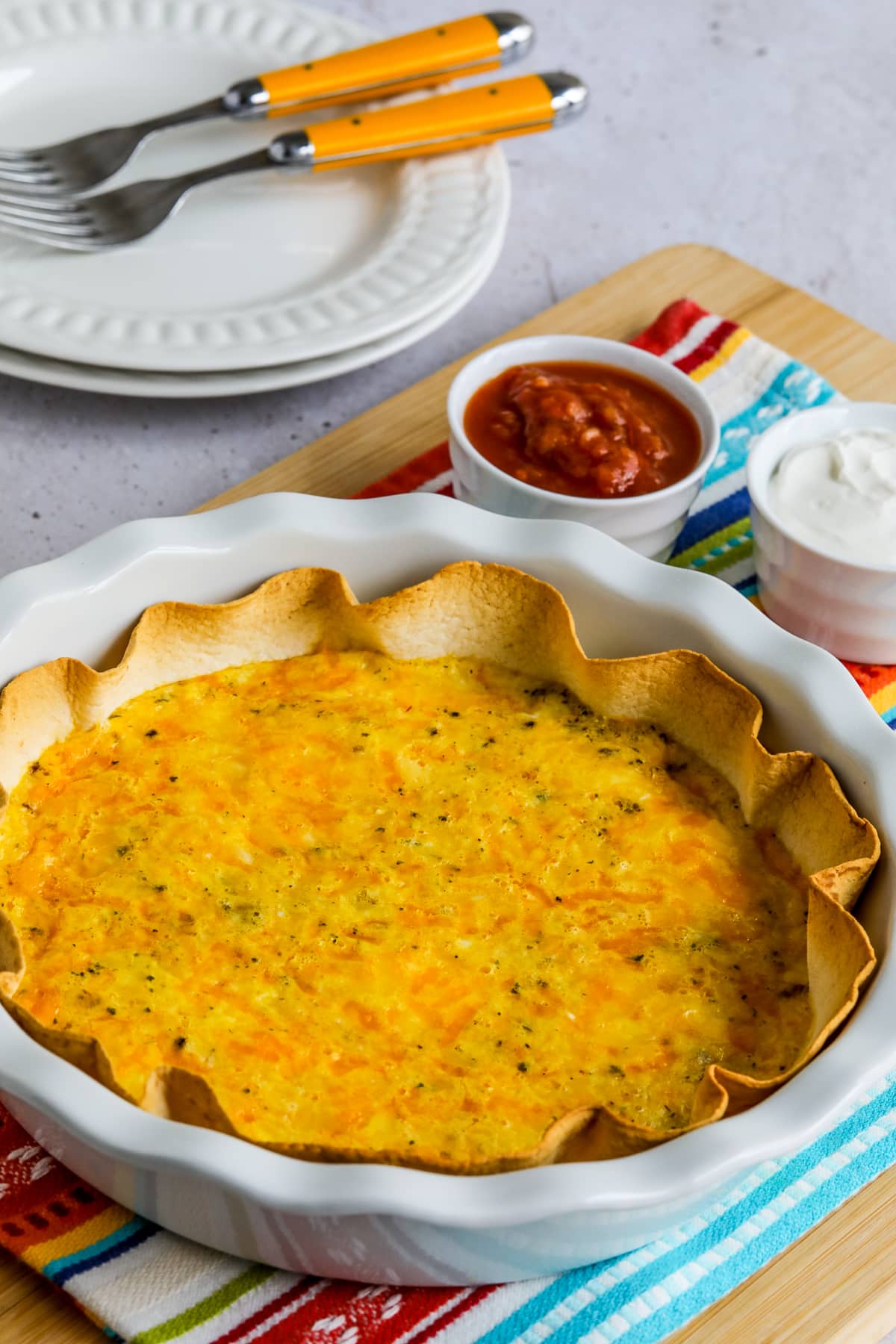 Tortilla Egg Bake shown in white pie dish with sour cream and salsa on the side and plates/forks in back.