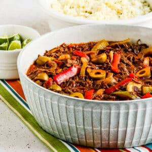 Square image of Ropa Vieja shown in serving bowl with limes and side dish.