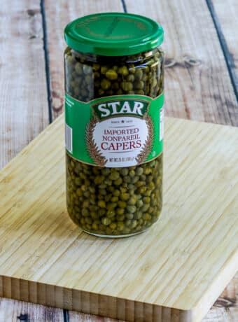 Square image of jar of capers on cutting board.