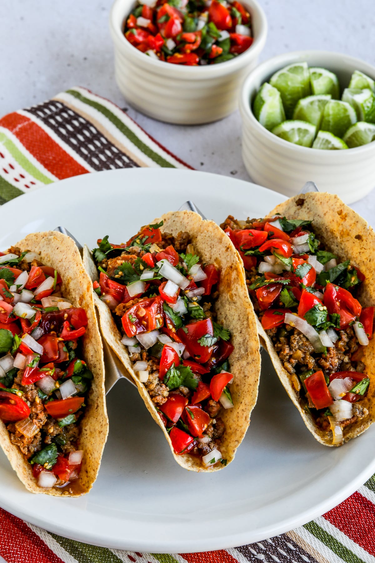 Three Beef and Chorizo Tacos shown in taco holder with limes and pico de gallo in back.