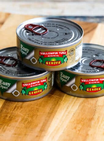 Square image for Genova Tuna in Olive Oil, showing three cans of tuna on cutting board.